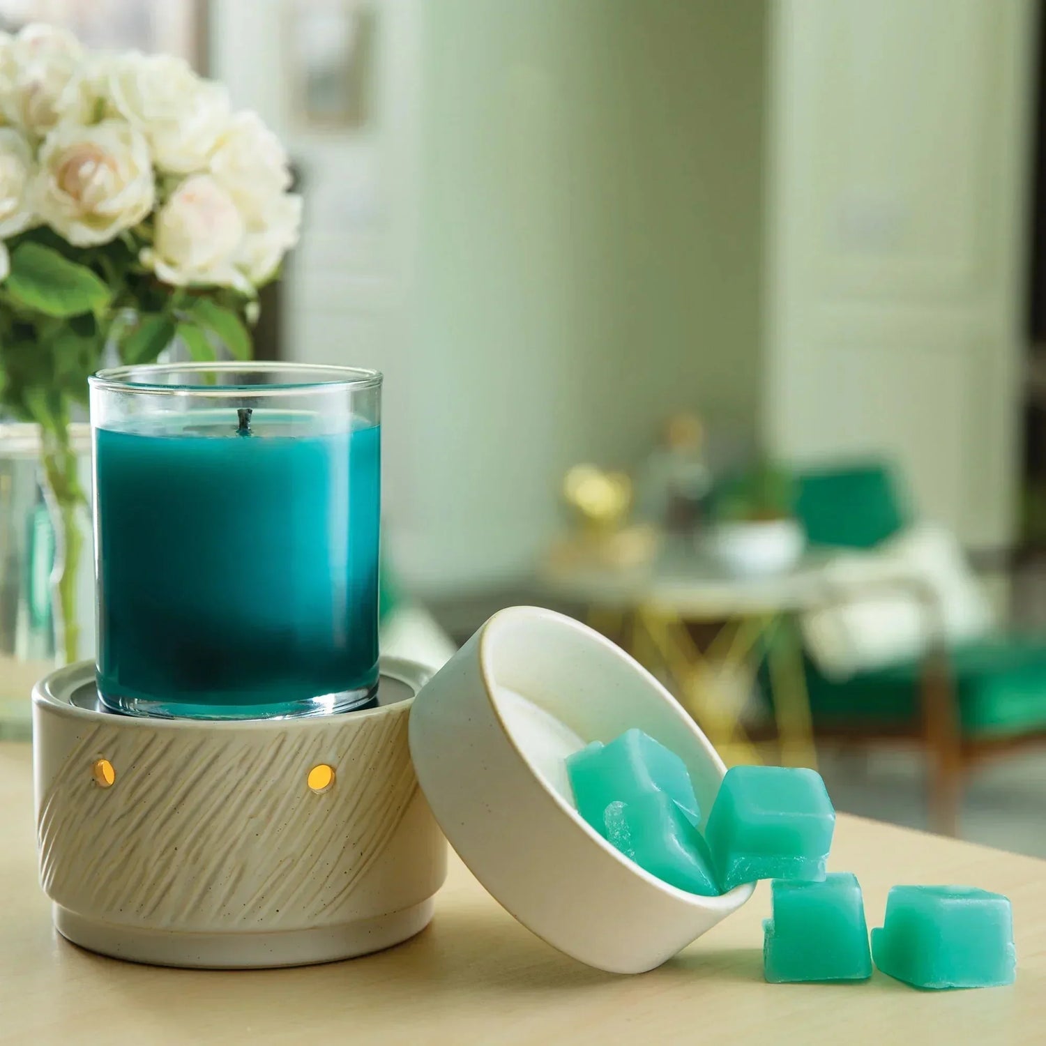 A teal candle sits on a ceramic warmer next to a bowl containing teal wax melts. A vase of white flowers and green furniture are blurred in the background.