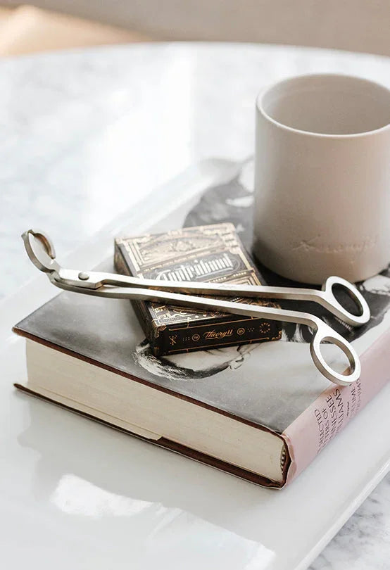 A pair of silver wick trimmers, a decorative matchbox, and a beige mug rest on top of a hardcover book on a white tray.