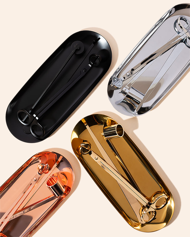 Four Makesy® Candle Care Kits in black, silver, rose gold, and gold are neatly arranged on oval trays. Each kit includes scissors, a wick trimmer, and other tools displayed on a light background.
