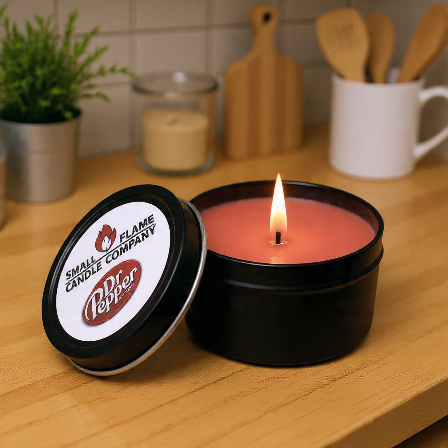 A lit Dr Pepper Candles and Melts red candle in a black tin from Small Flame Candle Company sits on a kitchen counter by jars, utensils, a mug, and a plant. Made with coconut soy wax and natural fragrance oils for an inviting aroma.