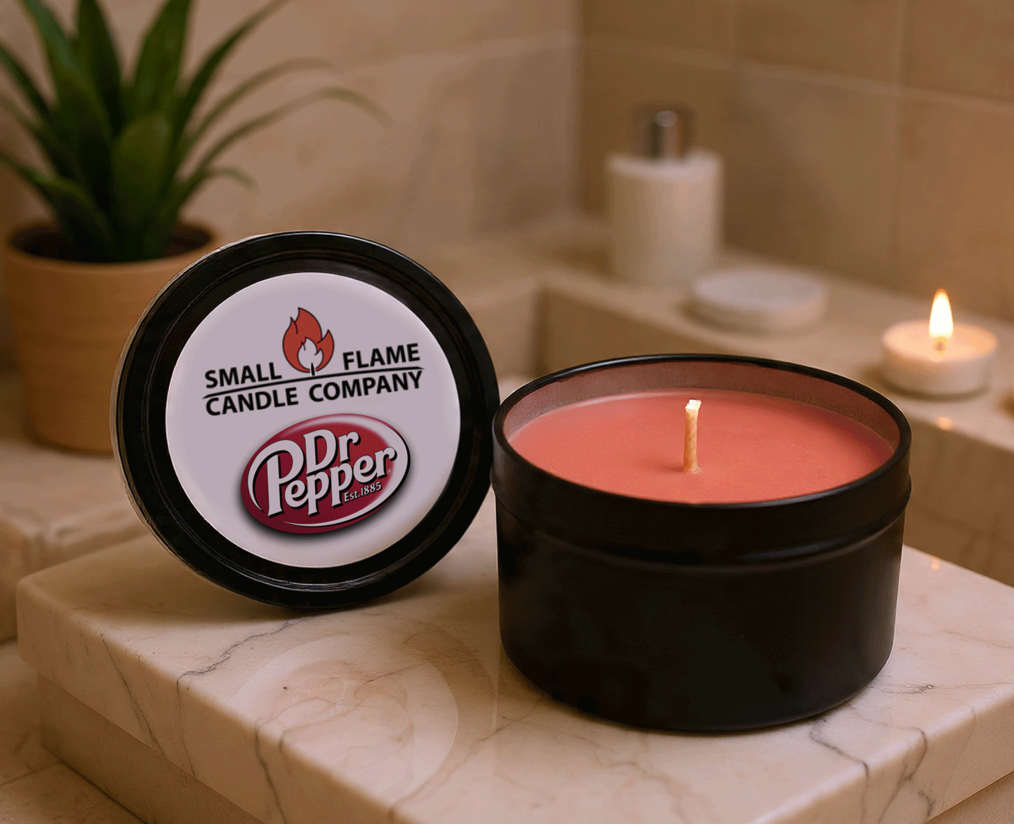 A lit reddish Dr Pepper Candles and Melts coconut soy wax candle sits in a black tin on a bathroom counter. The lid features the product name, while a green plant and another candle are softly blurred in the background.