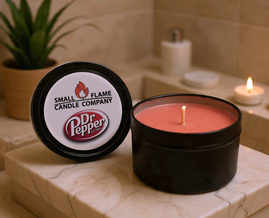 A lit reddish Dr Pepper Candles and Melts coconut soy wax candle sits in a black tin on a bathroom counter. The lid features the product name, while a green plant and another candle are softly blurred in the background.