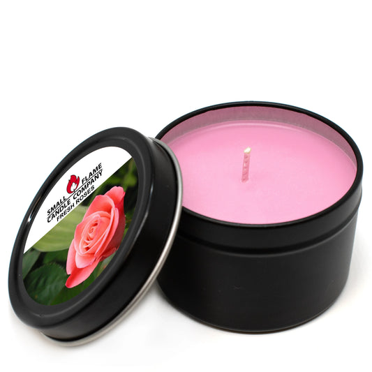 A pink, hand-poured Fresh Roses Candles and Melts candle sits in a black tin with the lid off. The lid displays a photo of a pink rose and text: SMALL FLAME CANDLE COMPANY FRESH ROSE, highlighting its fresh rose scent.