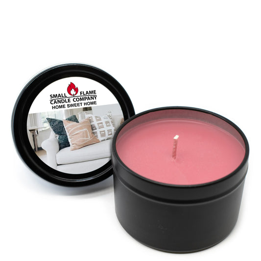 A pink Home Sweet Home Candles and Melts item sits in a black tin made with coconut soy wax and natural fragrance oils. The lid, showing the Small Flame Candle Company label and a white couch with pillows, rests beside it.