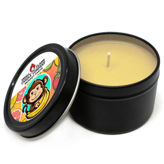 A black metal tin from Small Flame Candle Company holds a yellow Monkey Farts Candles and Melts coconut soy candle. The lid, beside it, shows a cartoon monkey with a banana, fruit images, and the brand’s label.