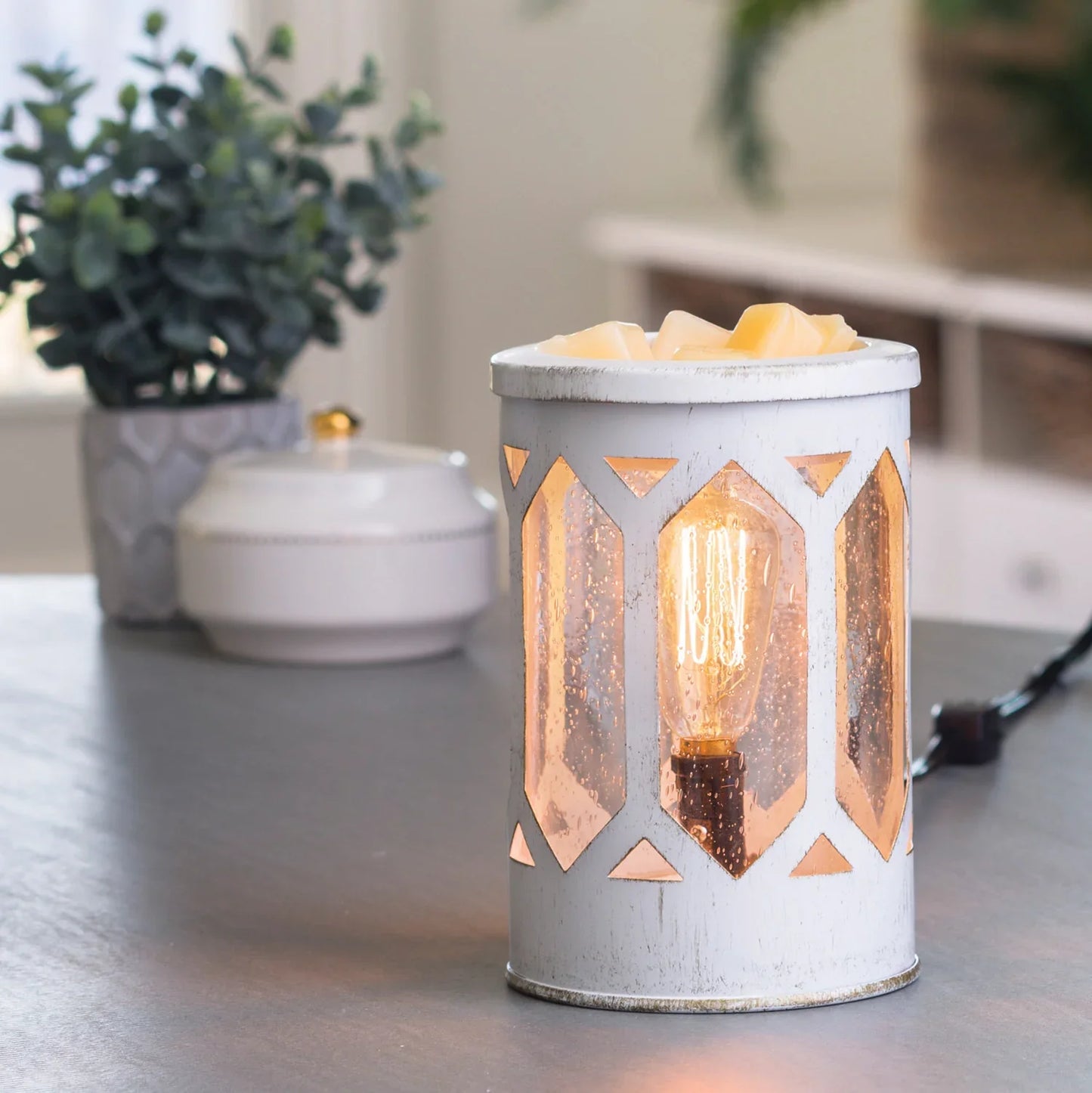 The Arbor Edison Bulb Illumination, a white lantern-style wax warmer with geometric cutouts, glows warmly on a gray tabletop. A potted plant and a white container with gold handle in the background bring vintage garden charm.