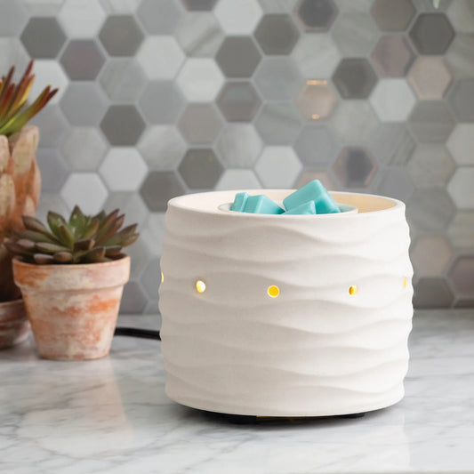 The Harmony Illuminaire Fan Warmer, a white ceramic piece with yellow light holes, sits on a marble counter beside potted succulents and blue wax melts, framed by a gray and white hexagon tile backsplash.