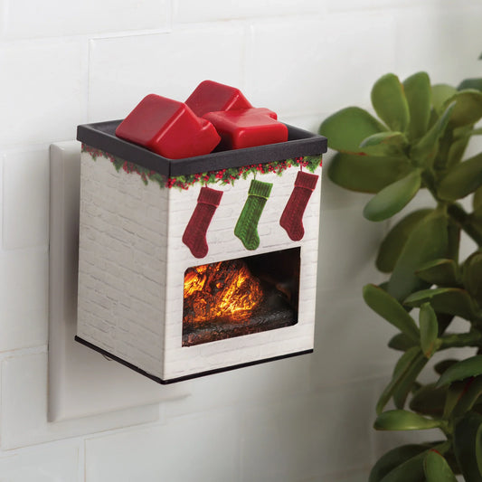 The Holiday Fireplace Pluggable, shaped like a whitewashed fireplace with festive details and red wax melts on top, plugs into an outlet near a green succulent to add charming seasonal decor.