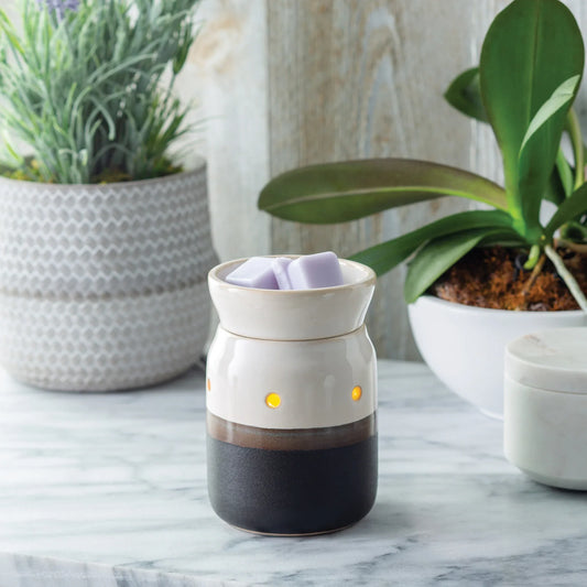 The Ivory & Iron Illumination wax warmer by Small Flame Candle Company glows with purple wax melts on a marble surface, surrounded by green potted plants in a bright room.