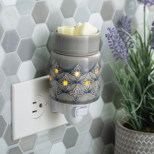 The Jasmine Pluggable wax warmer, featuring a grey crackle glaze and glowing lights, melts white wax while plugged into a wall outlet. It’s set against hexagonal tile with a potted lavender plant in the background.