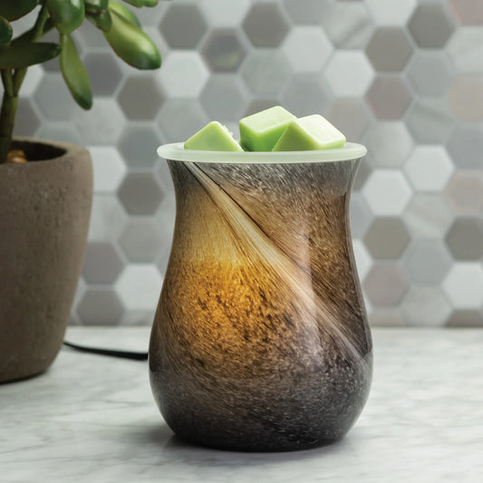 The Obsidian Illumination wax warmer by Small Flame Candle Company, with a glowing light, sits on a marble counter. Green wax melts rest on top beside a potted succulent; behind are marbled white swirls and gray-and-white hexagonal tiles.