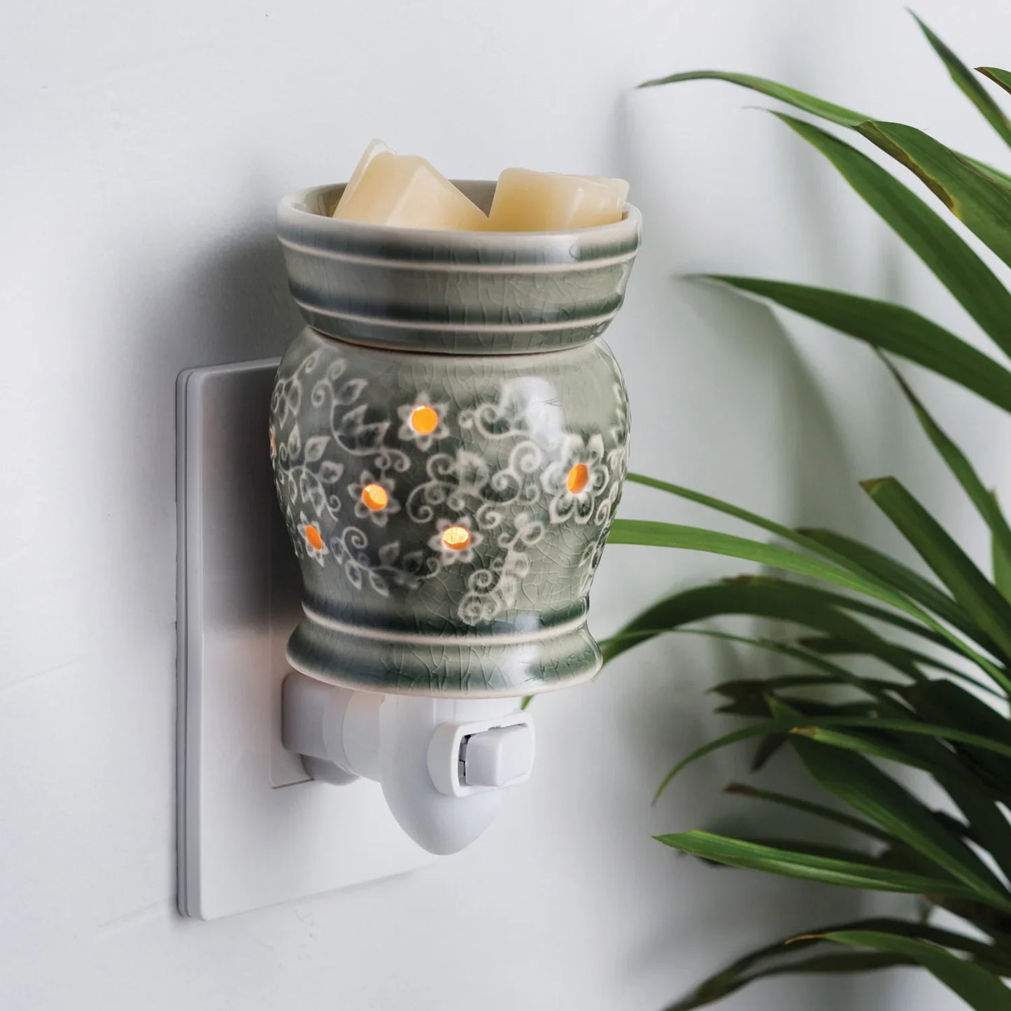 Ceramic electric wax melt warmer with floral design plugged into wall, surrounded by plant leaves