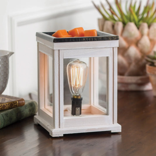 The Weathered White Edison Bulb Illumination, a square lantern-style wax warmer with vintage charm and a visible Edison bulb, holds orange wax melts on top. It sits on a wooden table near books, a bowl, and a succulent plant.